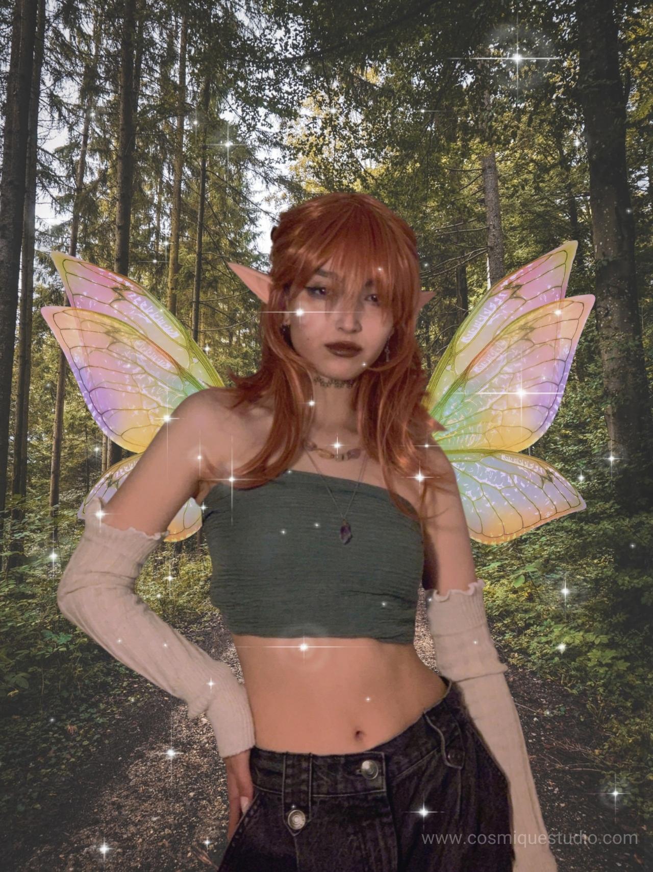 A girl who has fairy wings and elf ears standing in the forest wearing a green crop top.