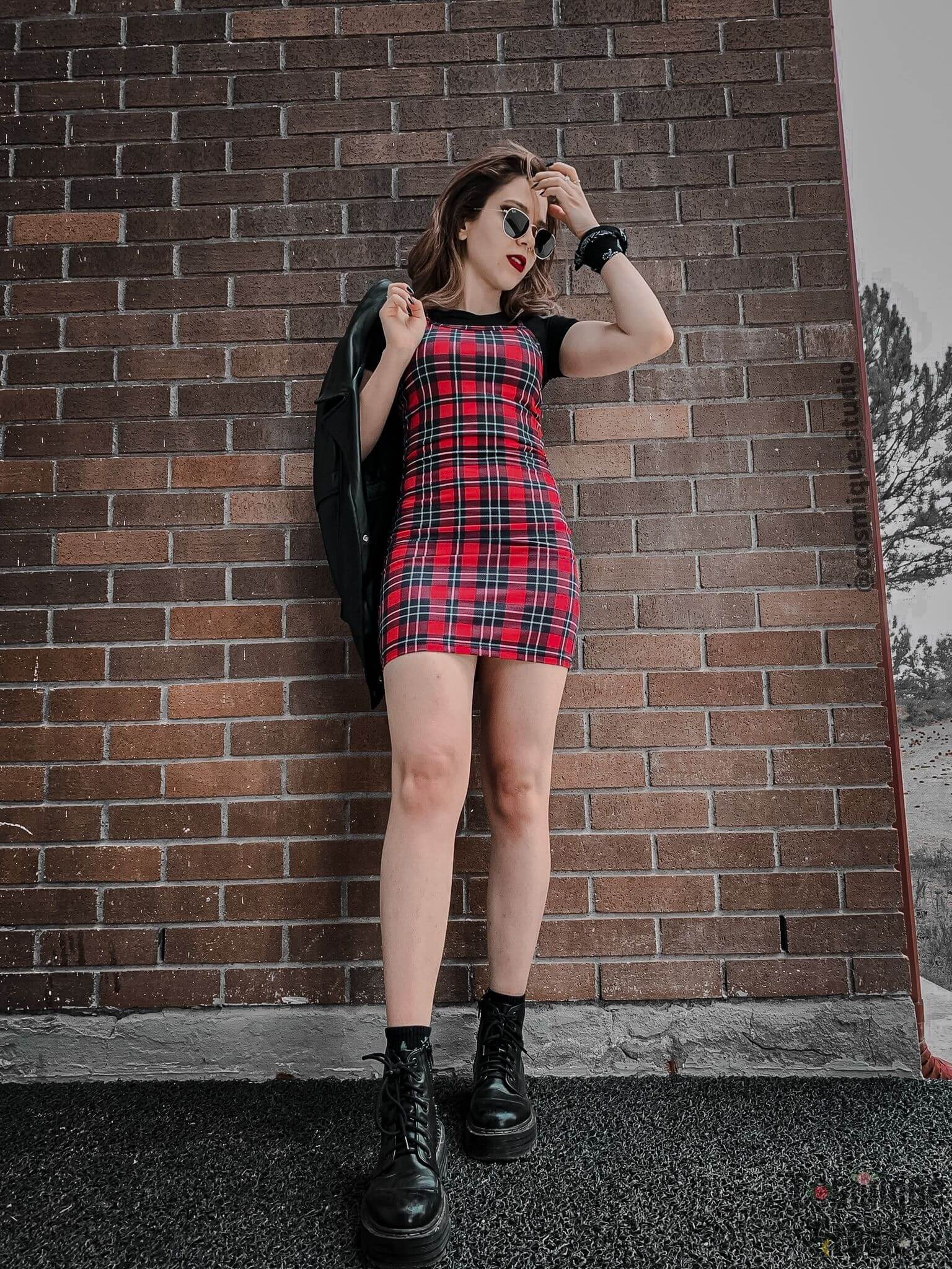 grunge girl on a tile wall, wearing plaid mini red dress combined with black platform boots, black leather jacket on her finger, and black sunglasses