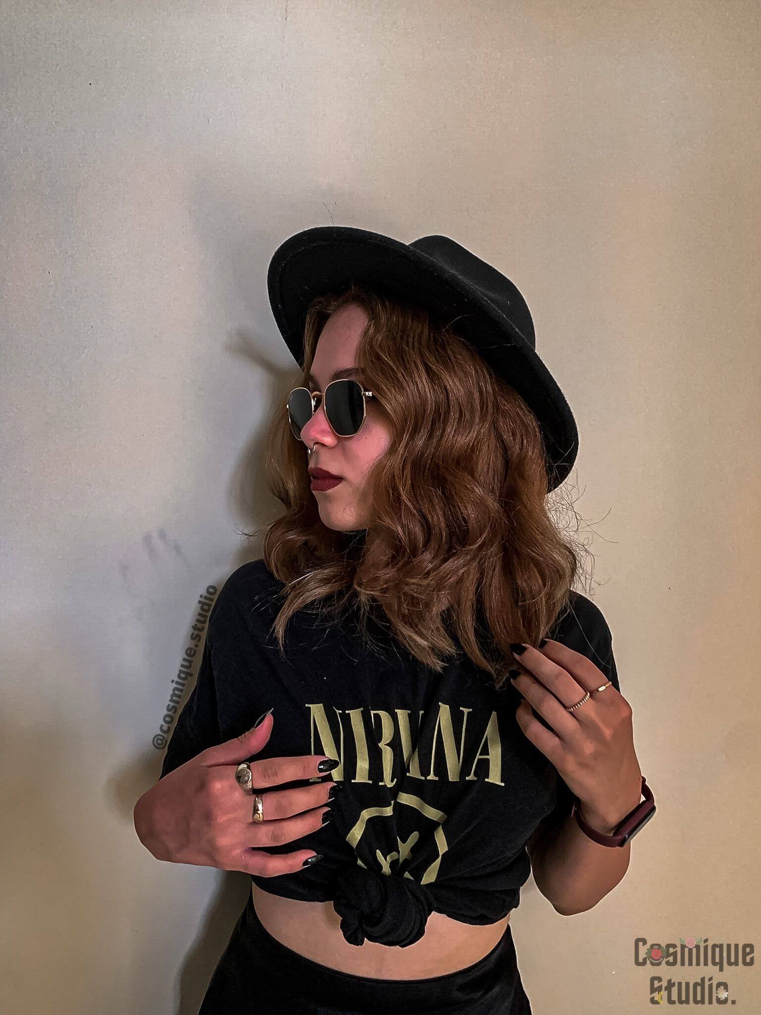 grunge girl in front of a wall wearing nirvana t-shirt combined with black fedora hat, gold rings, and sunglasses
