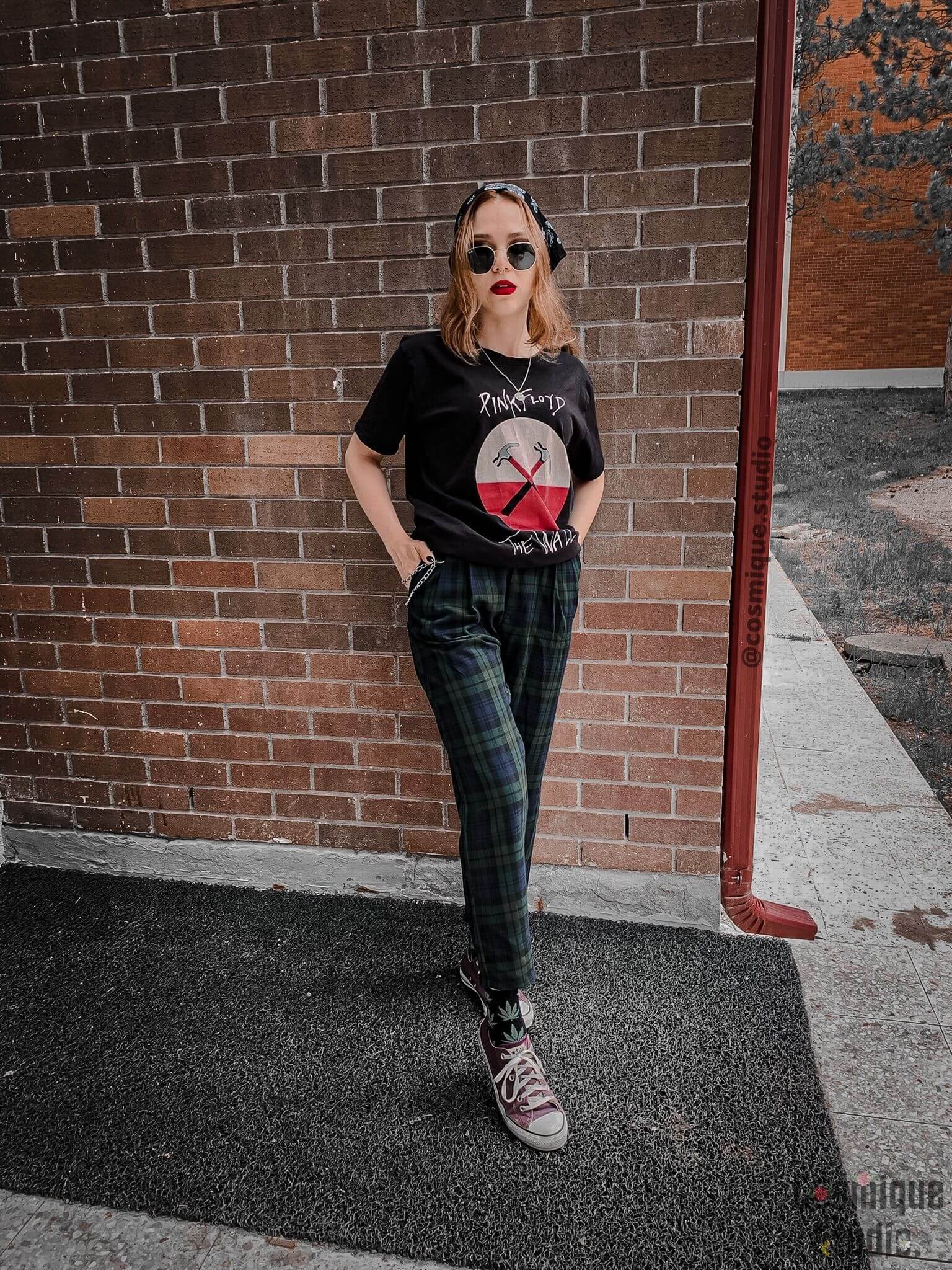 grunge girl outfit in front of a tile wall wearing pink floyd band t-shirt, plaid baggy pants combined with black bandana, weed socks, purple leather converse and sunglasses
